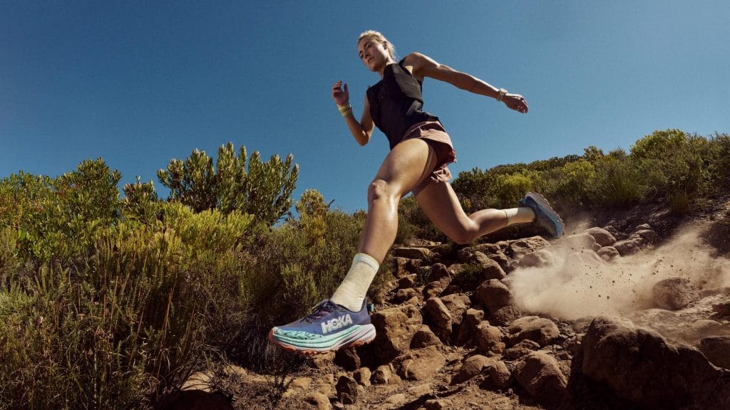 En kvinna springer nerför en stenig stig i ett soligt, grönt landskap. Hon bär träningskläder och blågrön-lila löparskor från Hoka, som virvlar upp damm när hon landar på marken.