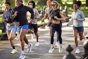 En grupp män och kvinnor springer tillsammans i en park. Det är sommar och de bär lätta träningskläder och löparskor från HOKA på fötterna. I bakgrunden syns grönt gräs och bänkar.