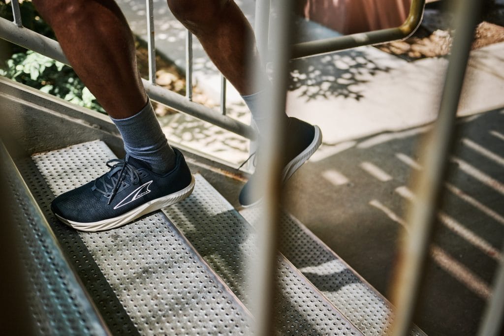 Person går uppför en metalltrappa i svarta löparskor med vit sula och grå strumpor. Bilden är tagen genom ett trappräcke i dagsljus.